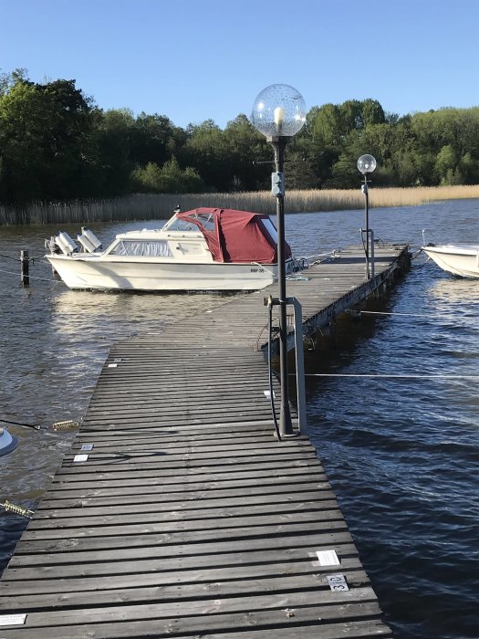 Motorbåt med röd kapell förtöjd vid en träbrygga på en sjö med träd i bakgrunden.