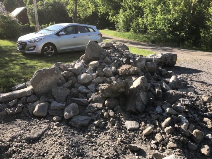 En hög med bortbilade betong- och stenklumpar på en grusig yta framför en bil och grönska.