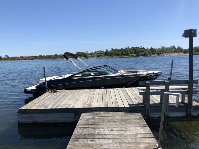 Motorbåt förtöjd vid en träbrygga på en solig dag, med blå himmel och lugnt vatten.