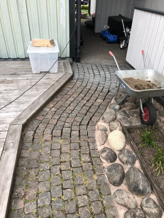 Renoverat stenlagt gångområde med kullerstenar och stenmjöl i en skottkärra, bredvid en trätrall och husvägg.