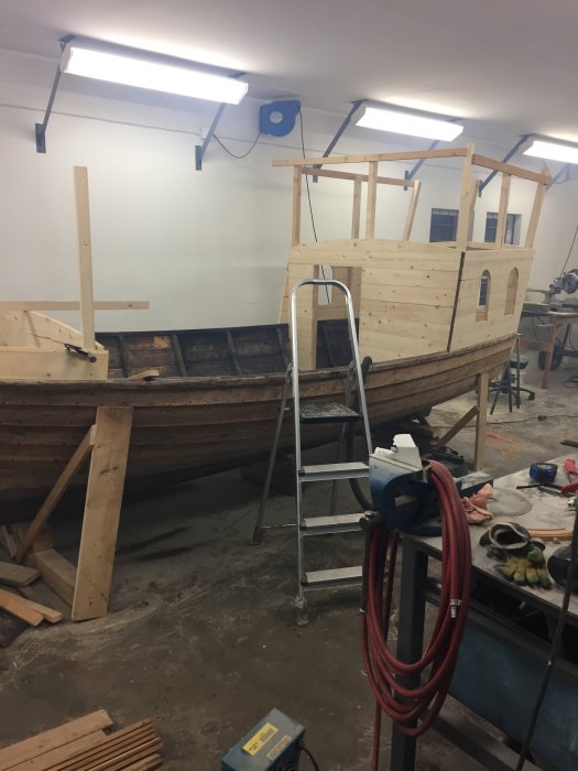 Träbåt under renovering i verkstad med verktyg och material synliga.