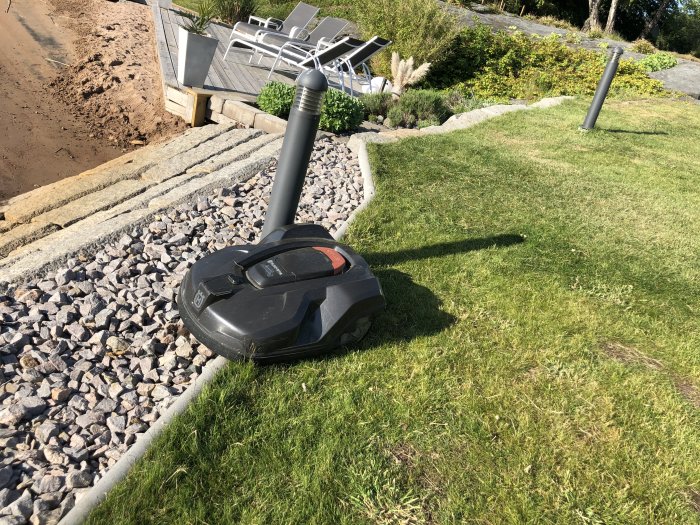 Gräsklippningsrobot nära kanten av en gräsmatta, på en stenbelagd väg mot en sandstrand.
