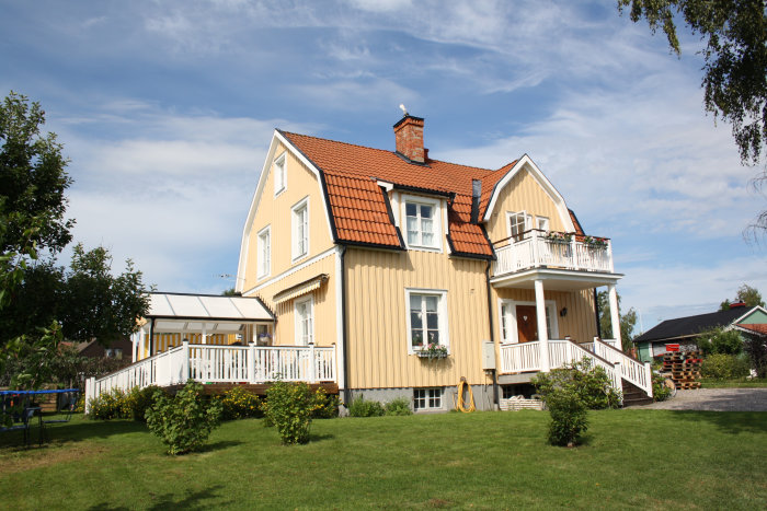 Gult tvåvånings trähus med tegeltak och veranda mot en klarblå himmel.