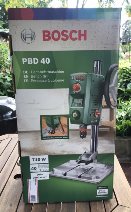 Förpackning av en Bosch PBD 40 bänkborrmaskin på ett träbord ute.