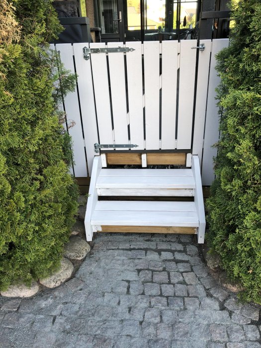 Ny målad vit tryckimpregnerad altantrappa mellan gröna buskar och stenläggning.
