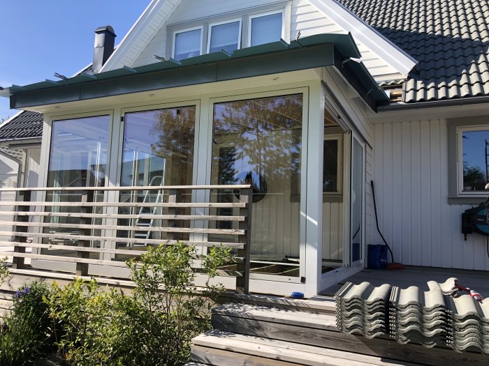 Ett halvfärdigt uterum med klart plåttak och nyinstallerade glaspartier mot en vitmålad husvägg.