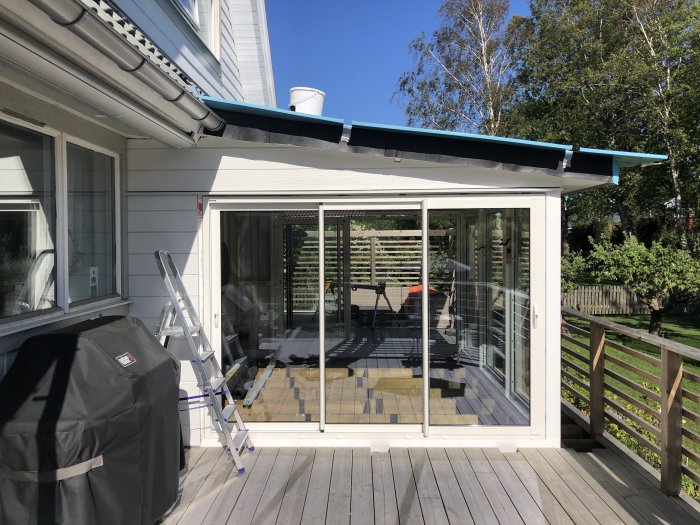 Nybyggt uterum med plåttak och glaspartier, intill en träveranda och vit husfasad under konstruktion.
