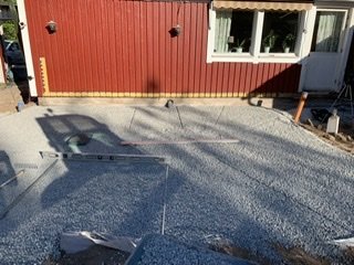 Byggarbetsplats med ojämnt utlagt grusmaterial 8/16 bredvid en röd husvägg, synlig skugga och vattenrör.