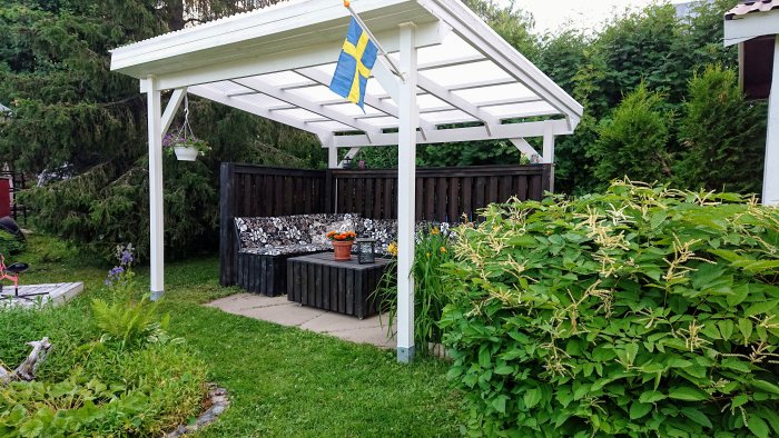 Trädgård med vit pergola, hembyggd soffa med dynor och kuddar, omgiven av grönska och en svensk flagga.