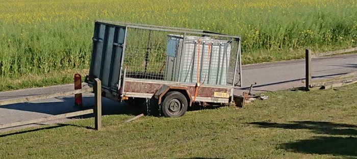 Övergivet släp med IBC-tank nära skadat staket vid vägkant, fält i bakgrunden.