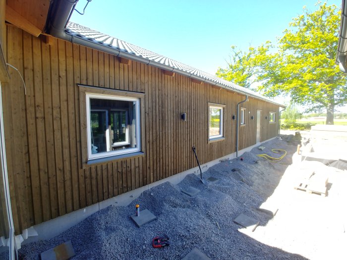 Hus med träpanel och fönster under markarbeten med grus och osatta plattor på tomten.