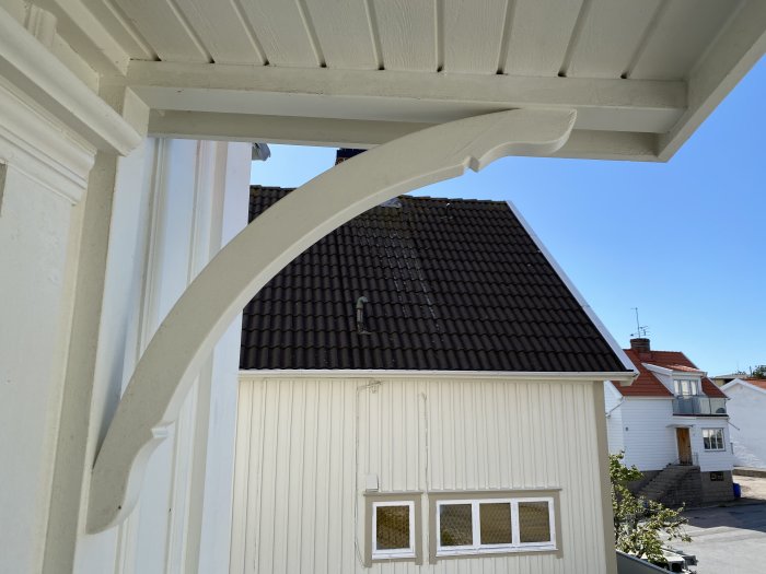 Dekorativt vitmålat trästöd monterat på entretak framför ett hus, med en bakgrund av tak och himmel.