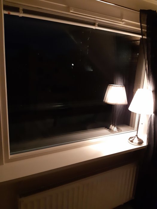 Stängt PVC-fönster i ett sovrum med bordslampa och gardiner vid nattetid.