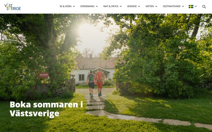 Två personer vandrar på en stig mot en lantlig stugby i soligt sommarväder.