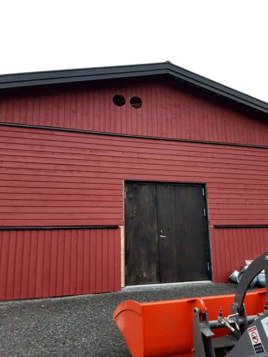 Röd träfasad på stall med stående och liggande dubbelfaspanel, svart dörr och två runda fönster.