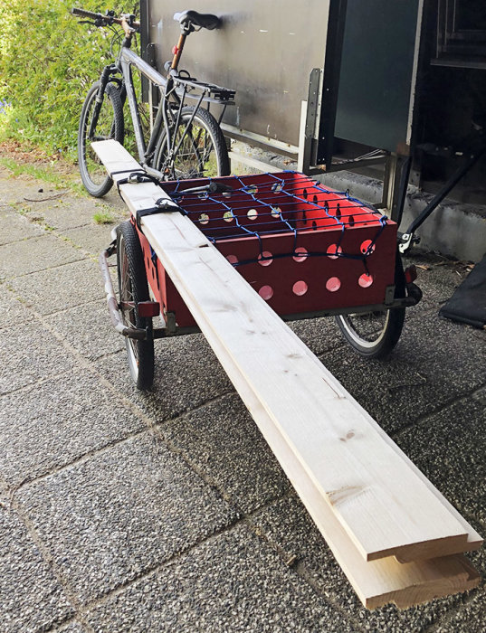 En ombyggd röd cykelvagn med träplankor säkrade ovanpå, parkerad framför en cykel och en släpvagn.