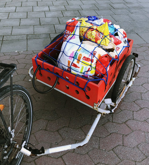 Cykelvagn fullastad med varor säkrade med blå nät, parkerad på stenbelagd mark.