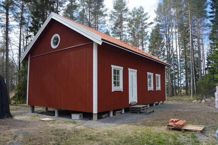 Rödmålat trähus med vitt fönsterfoder och ett runt loftfönster, gjutna plintar och byggmaterial i förgrunden.