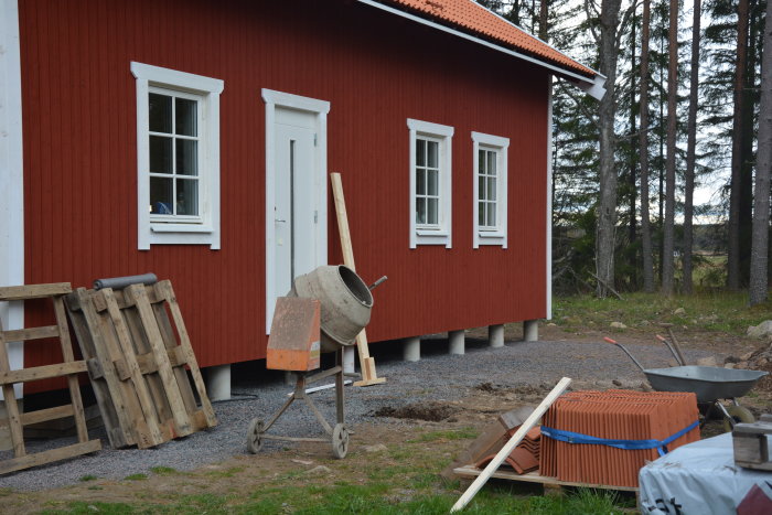 Renoveringsprojekt av rödmålad stuga, vit fönsterfoder och byggmaterial med betongblandare framför.