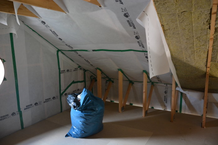 Takstolar och isolering i ett loft under renovering, med ångspärrstejp och skyddsplast synliga.