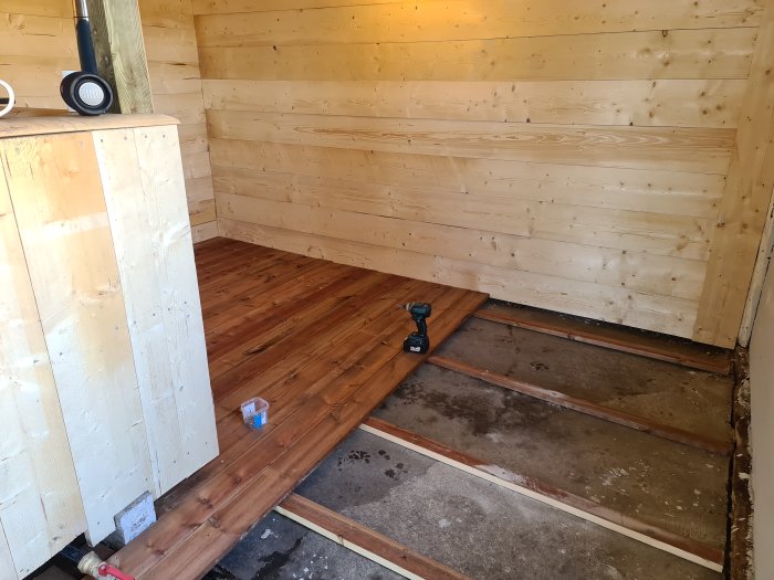 Inredning av ett under konstruktion badhus med träpaneler, trägolv och en borrskruvdragare på golvet.