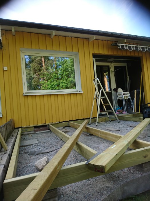 Pågående konstruktion av ett trädäck med ramverk av reglar framför en gul husvägg med ett fönster och öppen dörr.