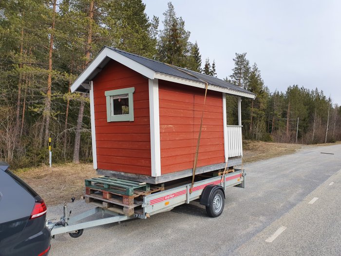 Rödmålat litet hus på en släpvagn intill en bil parkerad vid en landsväg.