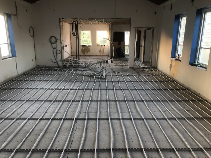 Ett golv under renovering med nyinstallerad golvvärme bestående av slangar fästa i skenor på betongunderlag.