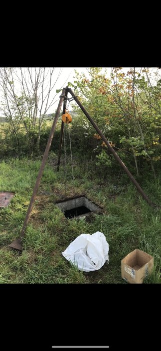 Manuell lyftställning av metall med kättingtelfer över öppen brunn i ett gräsfält.