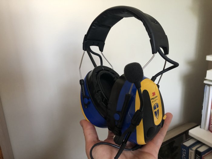 Hörselkåpor med gelfyllda öronkuddar, blåtand och bom-mikrofon hålls upp av en hand.