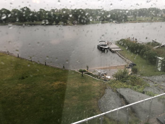 Utsikt från ett fönster under regnväder över en gräsmatta som leder till en brygga med en båt, omgiven av en sjö.