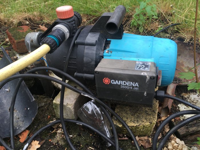 En Gardena 3500/4 Jet vattenpump med anslutna sladdar och slangar, belägen utomhus bland sten och vegetation.