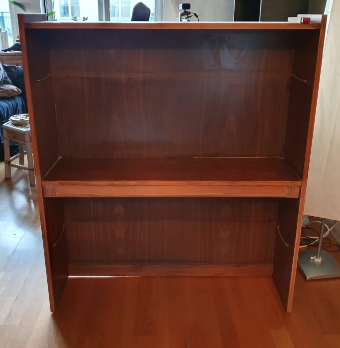 Tom gammal 60-talsbokhylla i massivt trä med mörk rödbrun betsning, placerad på ett trägolv i ett rum under renovering.