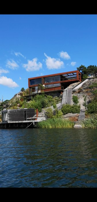 Modern arkitekthuset med stora fönster och trädetaljer, placerat på klippa vid vattenkanten.