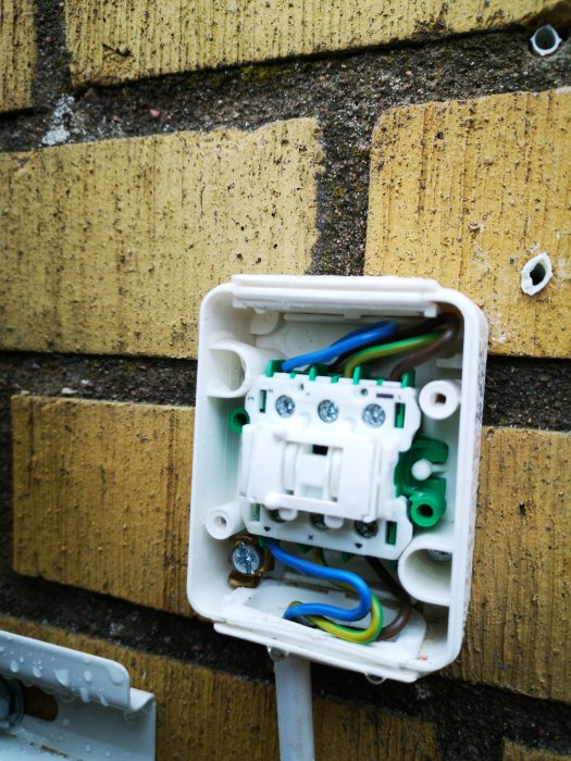 Öppen elektrisk anslutningsdosa monterad på en tegelvägg med synliga ledningar och kopplingar.