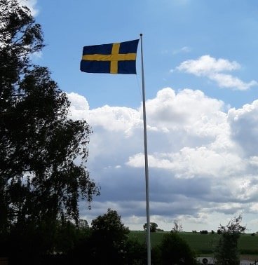 Svensk flagga i topp på en hög flaggstång mot molnig himmel.
