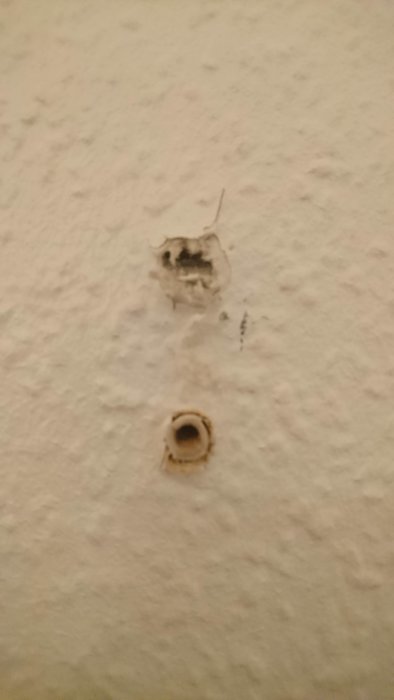 Ett större hål i en betongvägg ovanför ett rörformat hål vid ett eluttag, båda tecken på väggskador.