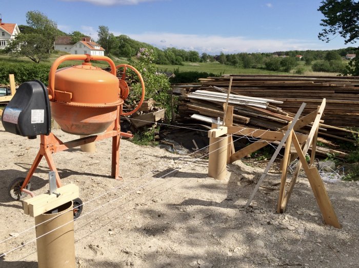 Byggplats med gräsmark, plintformar i papprör och en betongblandare, redo för gjutning.