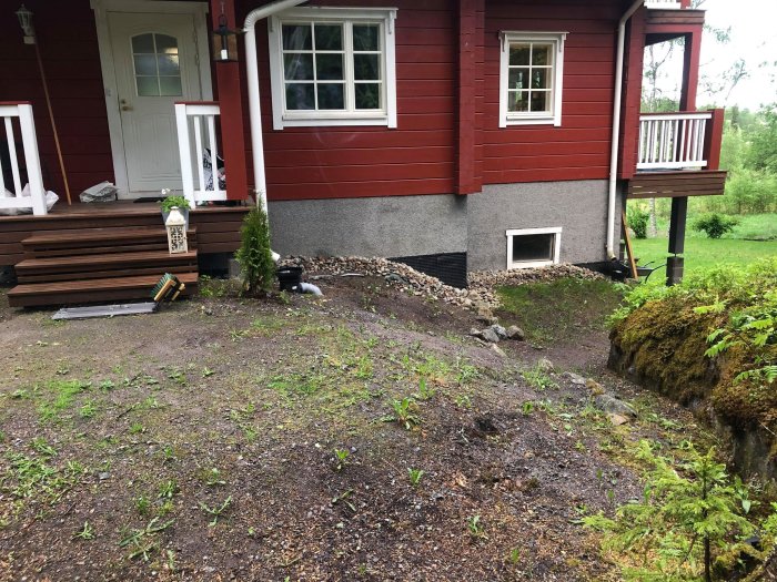 Rödmålat hus med trappa och balkong, oplanerad gård med gräs och grus, redo för landskapsdesign.