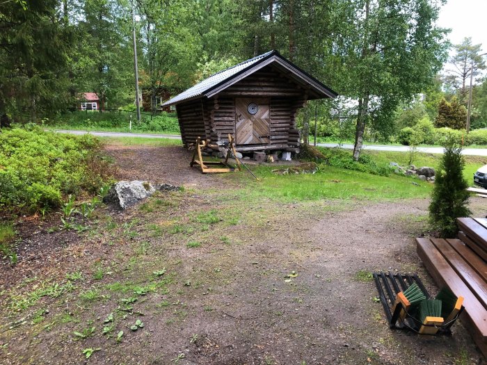 Gård med gräs och en grusgång som leder mot en trätrappa och en timmerstuga med grönska runtom.