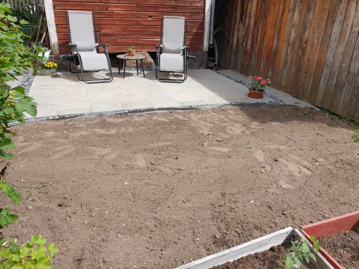 Ny anlagd uteplats med två solstolar och ett runt bord, omgiven av skifferkross, angränsande obehandlad jordyta.