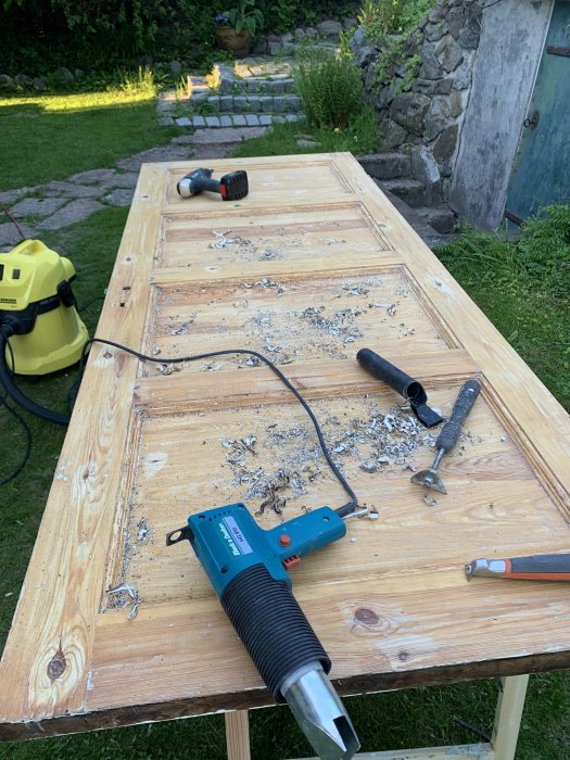 Nyrenoverad källardörr på ett arbetsbord med skrapor och slipverktyg, spån och målardetaljer i en trädgårdsmiljö.