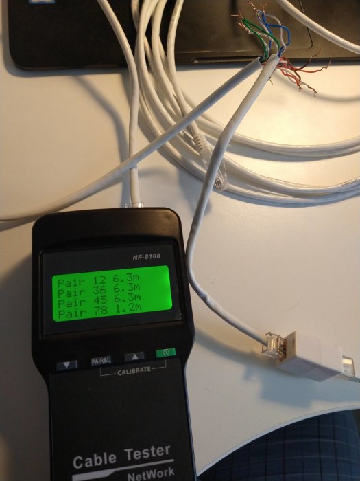 Avancerad nätverkskabeltestare med display visande par och längdmätning, oansenliga kablar i bakgrunden.