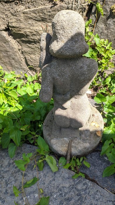 En stenstaty av en dam i betong med utstickande stålrör, omgiven av gröna bladverk på grå stenplattor.
