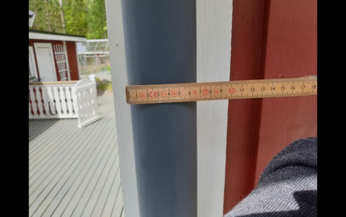 Mätning av ett grått 60 mm stuprör med en trälinjal på en veranda med röda stolpar.