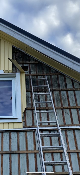 Äldre träfasad med tecken på slitage synlig efter borttagen plastpanel, med stegar lutade mot väggen.