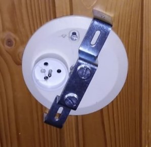 Vit väggplatta för elluttag med en påmonterad metallbygel för en IKEA Tross lampa.