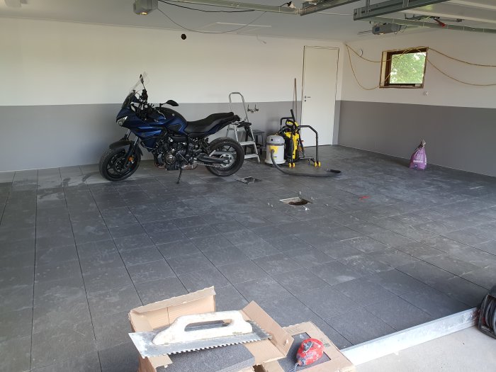 Nästan färdiglagt garagegolv med grå stenplattor, motorcykel, verktyg och renoveringsmaterial.