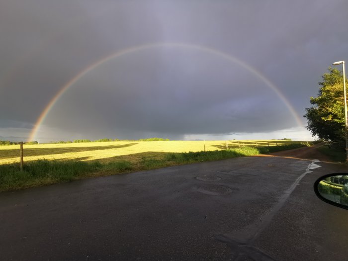 Hel regnbåge över en landsväg och fält, med mörka moln i bakgrunden och solsken i förgrunden.