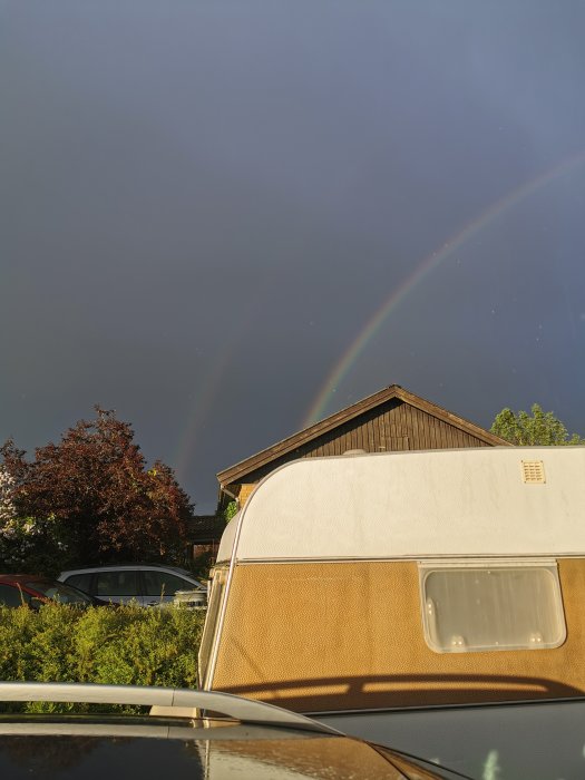 Dubbel regnbåge ovanför en husvagn och hus i ett bostadsområde efter regn.